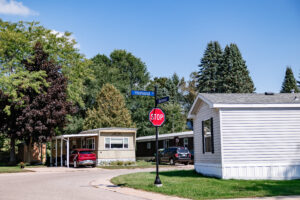 A stop sign stands at the corner of Highland and Belmont in Lakeview Village, a residential area with mobile homes and parked cars under a clear blue sky.