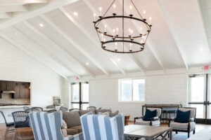 A spacious, bright living room in Lakeview Village with vaulted white ceilings, a large black chandelier, striped and neutral furniture, and natural light from multiple windows and doors.